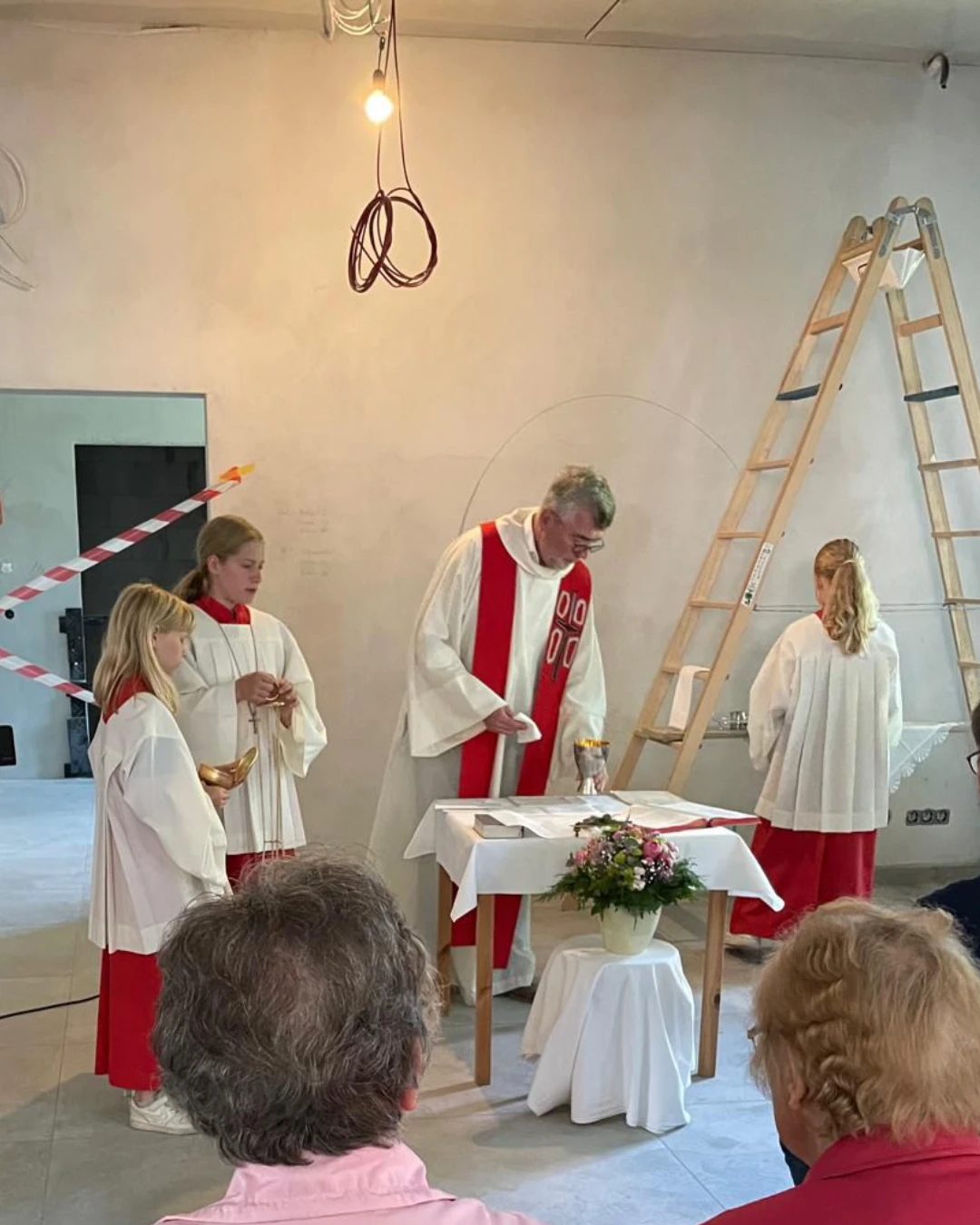 Pfarrer Bernd Strickmann feiert mit drei Messdienerinnen einen Gottesdienst auf der Baustelle im alten Pastorat – provisorischer Altar, unverputzte Wände, offene Kabel und eine Leiter im Hintergrund machen den Rohbaucharakter sichtbar.
