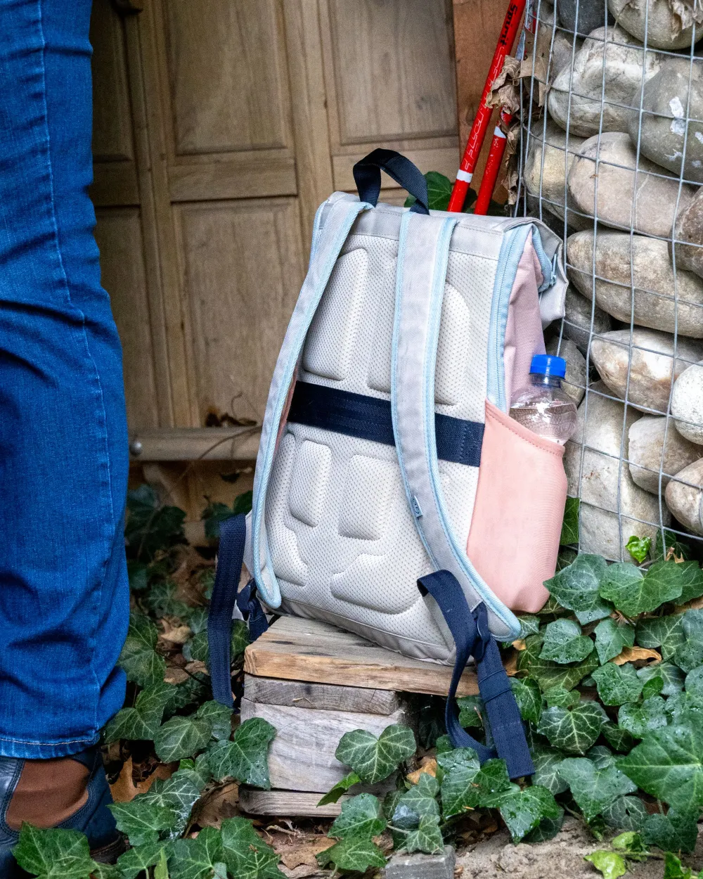 Ein hellgrauer Wanderrucksack mit hellblauen Trägern und rosa Seitentasche steht auf Holzklötzen am Boden, in der Seitentasche eine Plastikflasche mit Wasser; im Hintergrund Efeu, Steine und Holztür.